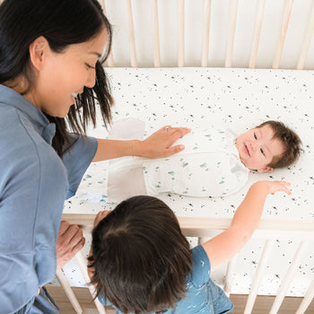 Mom and sister looking in to Lola Crib at baby swaddled on Ivory Galaxy Crib Sheet